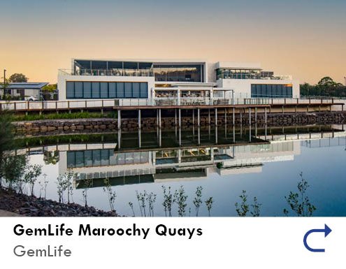 link to the GemLife Maroochy Quays project feature by the Australian National Construction Review. Image shows the club house of the retirement village from across a small body of water at dusk. the building is three levels and mostly white, with large boardwalk decks onto the lake and floor to ceiling glass windows.